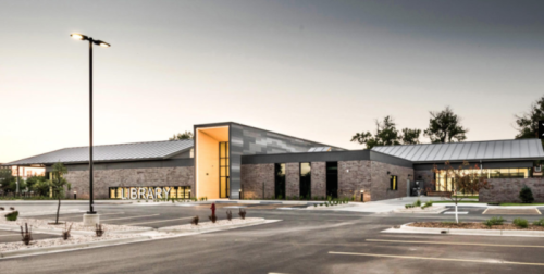Modern library building with large windows, stone facade, and illuminated entrance, set in a landscaped area.