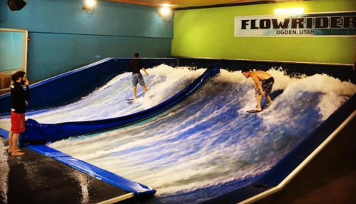 Two surfers ride artificial waves on a FlowRider in a recreational facility, with a spectator watching nearby.