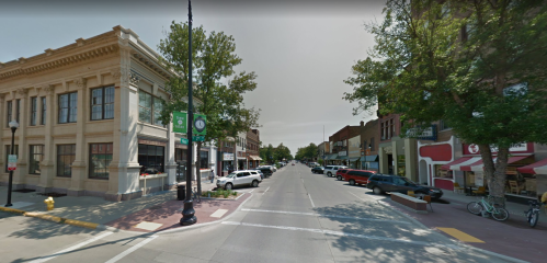 A view of a tree-lined street with shops and parked cars on either side, under a clear sky.