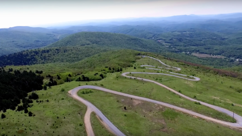 Aerial view of a winding road through lush green hills and mountains under a clear sky.