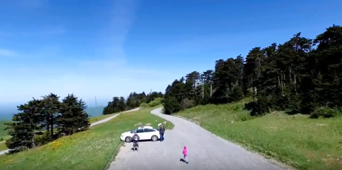 A scenic view of a winding road with a white car parked, surrounded by green hills and trees under a clear blue sky.