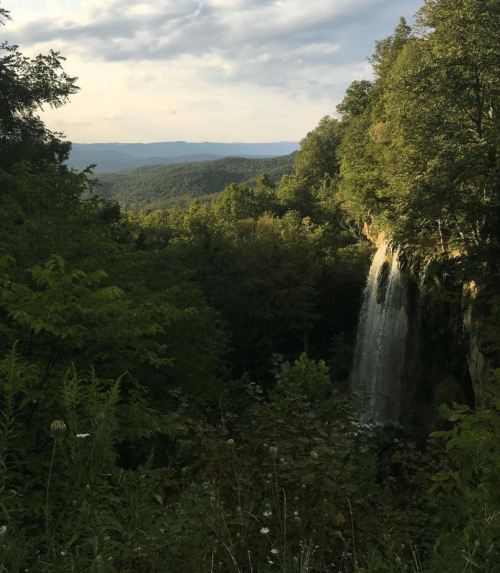 A scenic view of a waterfall surrounded by lush greenery and distant mountains under a partly cloudy sky.