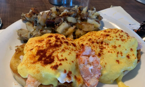 A plate of eggs Benedict topped with shrimp and hollandaise sauce, served with a side of seasoned potatoes.