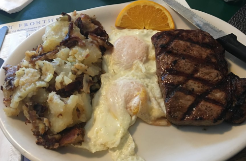 A plate with grilled steak, two fried eggs, and a side of crispy hash browns, garnished with an orange slice.