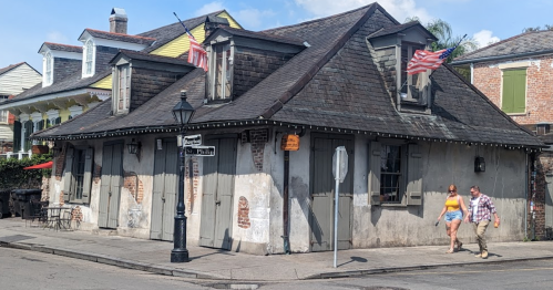 Two people walk past a historic building with American flags, featuring a weathered exterior and a streetlamp nearby.