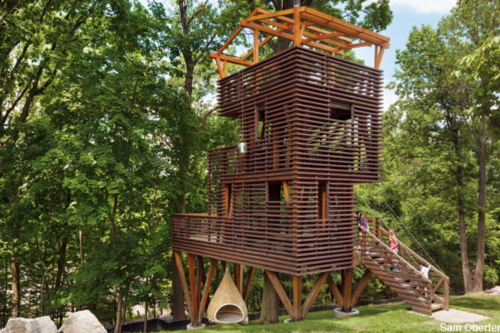 A modern treehouse with wooden slats, a balcony, and a hanging chair, surrounded by lush green trees.