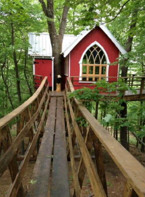 A whimsical red treehouse with a pointed roof and large windows, connected by a wooden walkway through lush greenery.