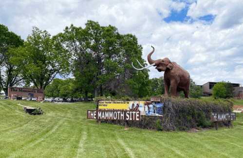 A large mammoth statue stands beside signs for "The Mammoth Site" on a grassy area under a cloudy sky.