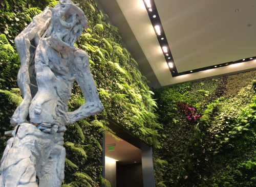 A tall, abstract white sculpture stands beside a lush green wall of ferns and plants in a modern interior space.