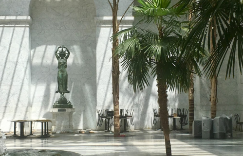 A marble atrium with a statue, palm trees, and empty tables under sunlight streaming through large windows.