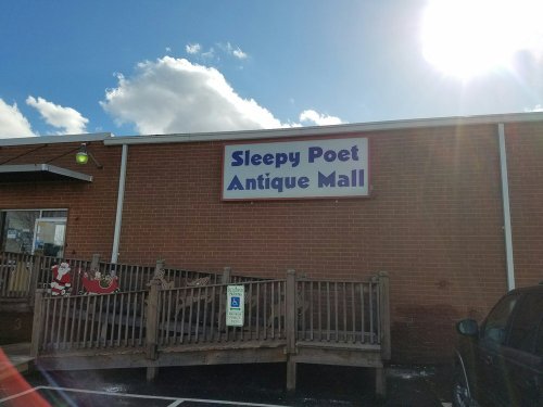 Sign for "Sleepy Poet Antique Mall" on a brick building, with a sunny sky and clouds in the background.