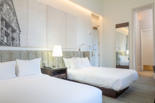 A modern hotel room featuring two beds, a lamp, and a large mirror, with light-colored walls and a stylish design.