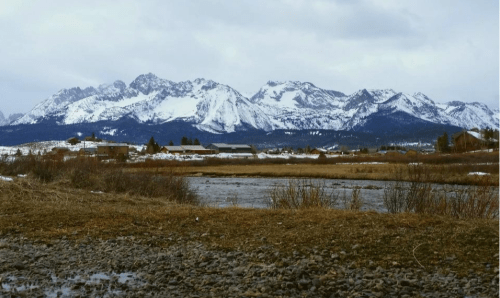 Snow-capped mountains rise above a grassy field and a calm river under a cloudy sky.