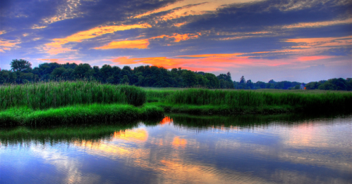 A serene landscape featuring a river reflecting a colorful sunset, surrounded by lush greenery and trees.