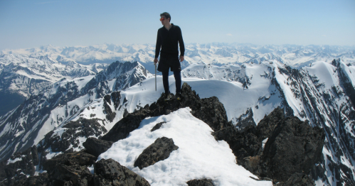 A person stands on a snowy mountain peak, surrounded by a panoramic view of snow-capped mountains under a clear blue sky.
