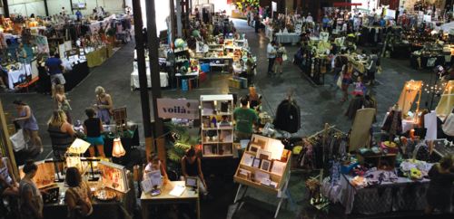 A bustling indoor market with various vendor booths, shoppers browsing, and colorful displays of crafts and goods.