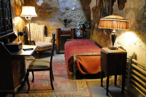 A dimly lit room with a bed, chair, desk, and lamps, featuring worn walls and a vintage rug.