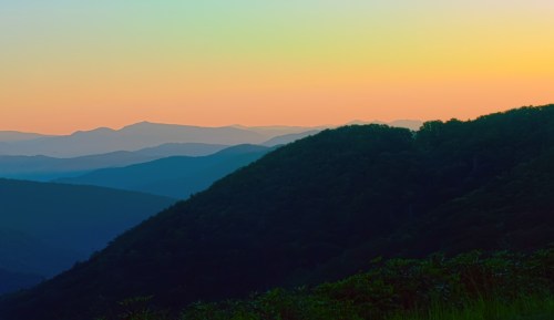 A serene landscape of rolling mountains at sunset, with soft hues of orange, blue, and purple in the sky.