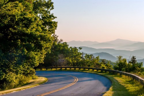 A winding road surrounded by lush greenery, with distant mountains and a soft sunrise in the background.