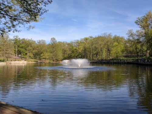A serene park scene featuring a pond with a fountain, surrounded by lush green trees under a clear blue sky.