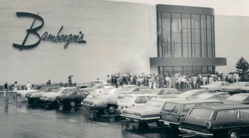 A crowd gathers outside Bamberger's department store on a rainy day, with parked cars in the foreground.