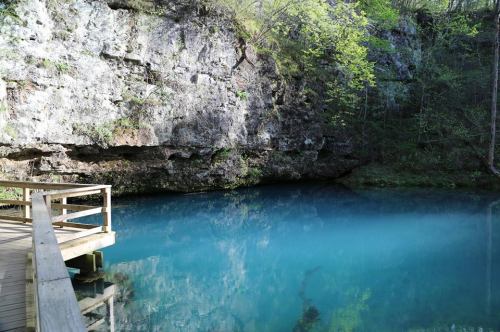 A serene blue pond surrounded by rocky cliffs and lush greenery, with a wooden walkway leading to the water's edge.