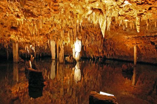 A dimly lit cave with stalactites hanging from the ceiling, reflecting in a calm underground pool of water.