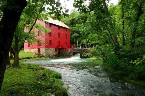 A red historic mill surrounded by lush green trees, with a flowing stream in front.
