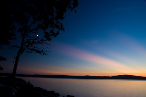 A serene lakeside scene at dusk, with a crescent moon and soft pastel colors in the sky.