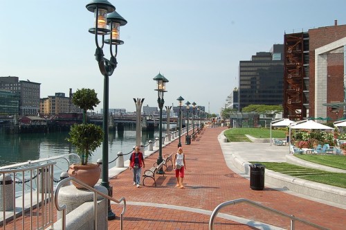 A waterfront walkway with people strolling, lined with lamps and greenery, overlooking a calm harbor.