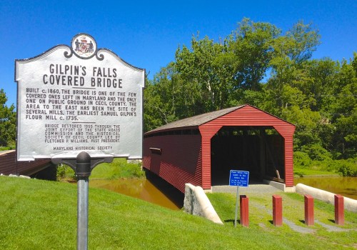A red covered bridge near a historical marker, surrounded by greenery and reflecting in the water.