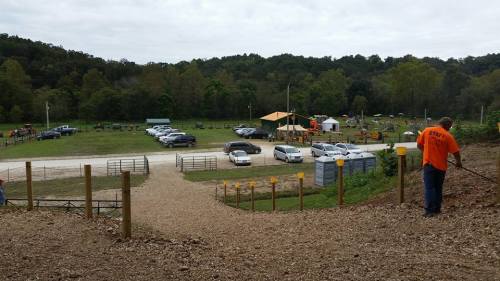 A gravel path leads to a parking area with cars, surrounded by trees and a distant activity area. A person is working nearby.