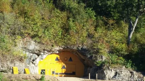 A yellow bunker entrance with warning symbols, set against a rocky hillside and surrounded by trees.