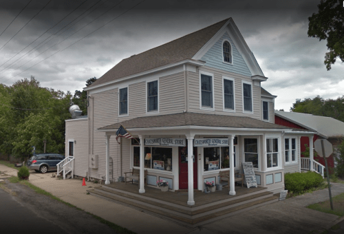 A two-story general store with a porch, featuring large windows and an American flag, set in a rural area.