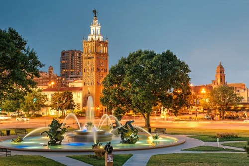 A city fountain with sculptures, illuminated at dusk, with a historic tower and buildings in the background.