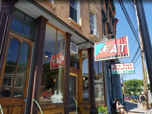 Exterior of a café with large windows and a sign reading "EAT Creekside Café," next to a pizzeria. Sunny day.