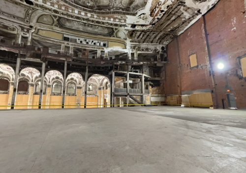 Interior of an abandoned theater, featuring peeling paint, exposed brick, and a large empty stage area.
