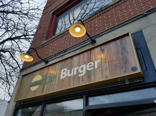 Sign for "Eden Burger" with wooden paneling and lights, mounted on a brick building. Tree branches visible above.