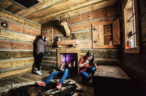 Three people in a rustic cabin, engaged in an escape room challenge, with a deer head mounted on the wall.