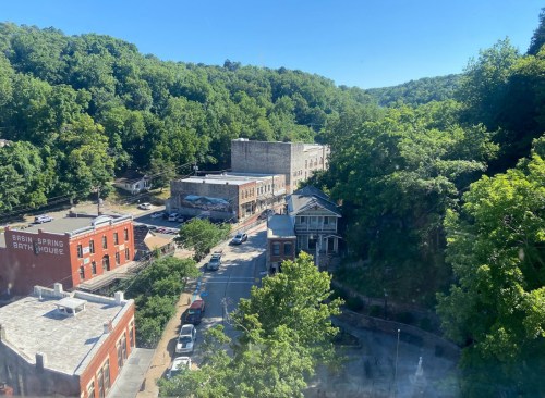 Aerial view of a small town surrounded by lush greenery, featuring historic buildings and a clear blue sky.