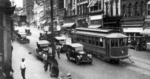 Historic street scene featuring vintage cars and a streetcar, bustling with pedestrians and storefronts.