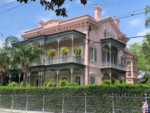 A large pink Victorian-style house with intricate ironwork balconies, surrounded by lush greenery and palm trees.