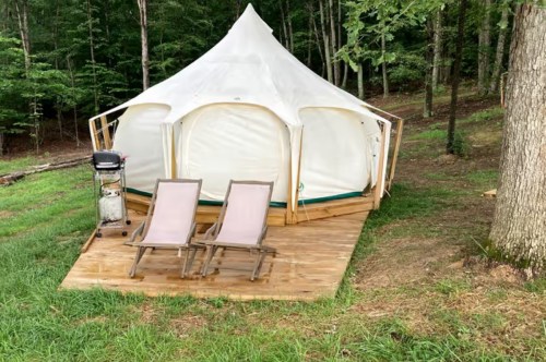 A white tent on a wooden deck in a forested area, with two lounge chairs and a grill nearby.