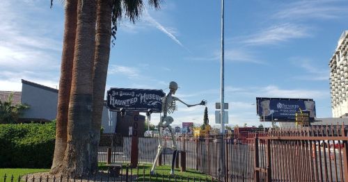 A skeleton figure stands outside a haunted museum, with palm trees and billboards in the background under a blue sky.