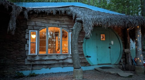 A cozy, rustic cabin with a thatched roof, round turquoise door, and warm light glowing from the windows.