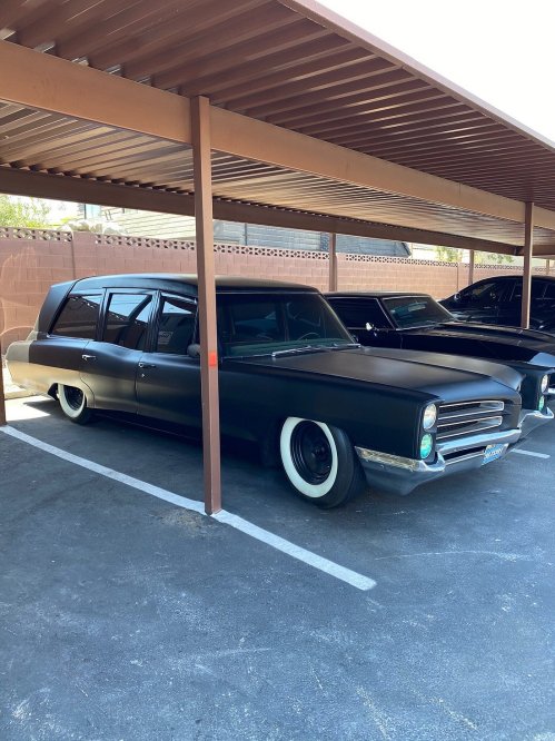 A vintage black car with whitewall tires parked under a carport, next to another dark vehicle.