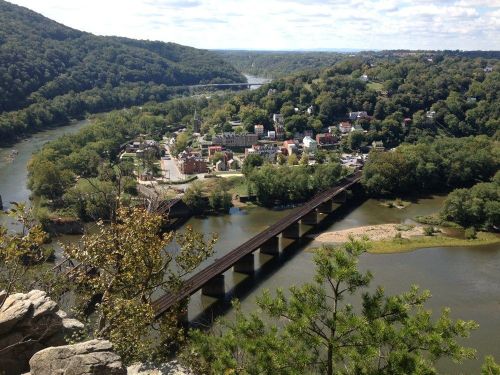 A scenic view of a river valley with a town and a bridge, surrounded by lush green hills under a blue sky.