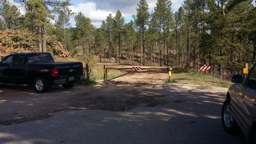 A dirt road entrance is blocked by a gate, with two parked vehicles nearby and trees in the background.