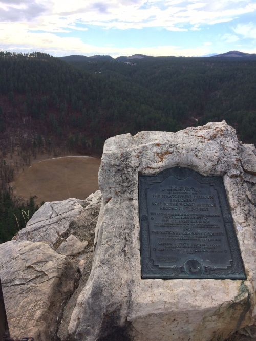 A plaque on a rocky outcrop overlooks a vast forested valley and distant mountains under a cloudy sky.