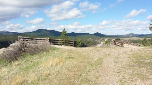 A scenic view of a grassy path leading to wooden gates, surrounded by mountains and a blue sky with clouds.